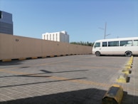 A nearly empty parking lot with a single white bus parked on the right side. The ground is paved with gray bricks, and there are yellow and black concrete parking stops. A beige wall runs along the side of the lot with a few round lights attached. In the background, there are a couple of modern buildings and some trees under a clear blue sky.