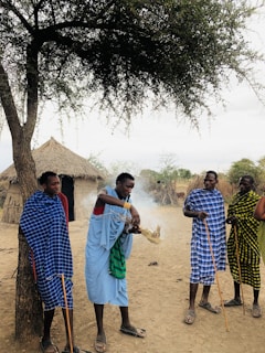A group of four individuals stands in a rural setting, with grass-covered huts in the background. Three of them hold long sticks, and one person is tending to a small fire that releases smoke. The individuals are wearing colorful, patterned fabrics, and a tree provides shade in the foreground.