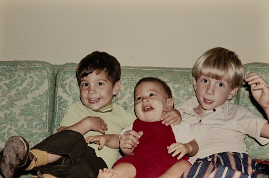 Three young children are sitting together on a patterned green couch. The child on the left is smiling widely, wearing a light green shirt and dark pants with yellow socks. In the middle, a toddler is wearing a red outfit, also smiling and appearing happy. The child on the right, with light blonde hair, is wearing a white shirt and colorful striped pants, looking towards the camera with a gentle smile.