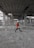 A person in a red jacket and white hard hat walks through a large, unfinished construction site. The area is filled with concrete columns, scattered debris, and construction materials. The ceiling exposes a grid structure, and rebar reinforces parts of the structure.