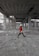 A person in a red jacket and white hard hat walks through a large, unfinished construction site. The area is filled with concrete columns, scattered debris, and construction materials. The ceiling exposes a grid structure, and rebar reinforces parts of the structure.