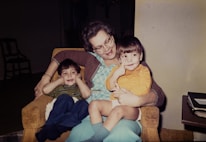 An older woman sits on an armchair with two young children on her lap. The woman is smiling and appears to be enjoying the moment with the children, who are also looking content. One child is dressed in a striped shirt and blue pants, while the other child is in a yellow top and light blue shorts. A stack of books or papers sits on a table nearby.