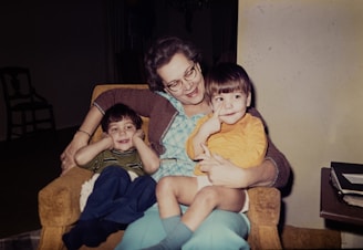 An older woman sits on an armchair with two young children on her lap. The woman is smiling and appears to be enjoying the moment with the children, who are also looking content. One child is dressed in a striped shirt and blue pants, while the other child is in a yellow top and light blue shorts. A stack of books or papers sits on a table nearby.