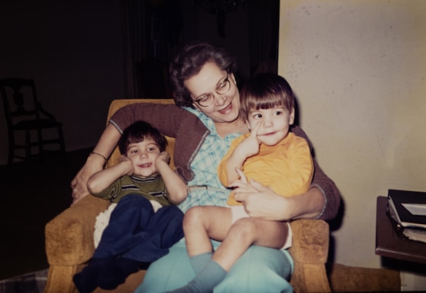 An older woman sits on an armchair with two young children on her lap. The woman is smiling and appears to be enjoying the moment with the children, who are also looking content. One child is dressed in a striped shirt and blue pants, while the other child is in a yellow top and light blue shorts. A stack of books or papers sits on a table nearby.