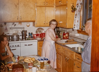 A vintage kitchen scene with wooden cabinets and a white stove. An elderly woman is standing at the counter, wearing a pink dress and apron, using a rolling pin near flour and baking items. A man with glasses looks at her, while another woman sits at the kitchen table, leaning her face on her hand. The table is set with various food items, including bananas, a cake, and a plate of bread. Decor items and cooking utensils are visible around the kitchen.