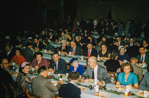 A large gathering of people seated at long tables covered with white tablecloths, engaged in conversation and enjoying a meal. The setting appears to be a banquet or formal event, with men in suits and ties and women in dresses. Plates with food and cups are visible on the tables, along with simple floral decorations. The crowded room is filled with a diverse group of attendees, indicating a lively social atmosphere.