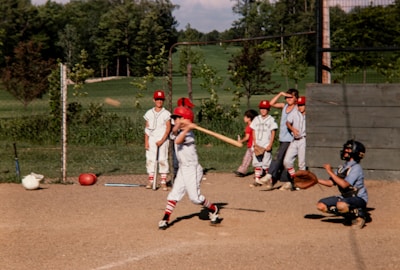 A group of young baseball players are in action on a sunny day. One player is swinging a bat, while another in catcher's gear crouches behind him. Several teammates, dressed in matching uniforms and red caps, stand by in the background, observing the game. The scene is set against a backdrop of lush greenery and a clear sky.