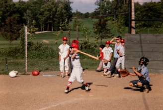 A group of young baseball players are in action on a sunny day. One player is swinging a bat, while another in catcher's gear crouches behind him. Several teammates, dressed in matching uniforms and red caps, stand by in the background, observing the game. The scene is set against a backdrop of lush greenery and a clear sky.