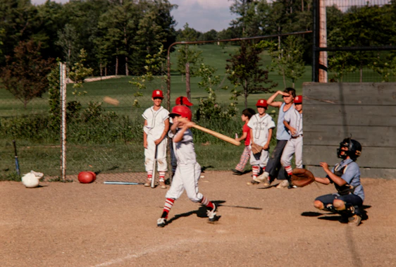 A vibrant action shot capturing a young player sliding into base with teammates cheering in the background under a clear blue sky.