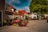 A quaint street features charming buildings with red-tiled roofs and vibrant flower arrangements. Colorful flags hang from the buildings, and large trees provide shade over the cobbled street. In the background, outdoor seating and a small cart add to the village-like atmosphere.
