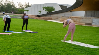 Three people are practicing yoga on mats in a lush green park. They are performing various poses and stretches, surrounded by modern architecture and greenery.