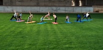 A group of people is practicing yoga on colorful mats spread across a lush green lawn under a concrete structure. The participants are performing a stretching pose and are dressed in casual athletic wear.