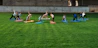 A group of people is practicing yoga on colorful mats spread across a lush green lawn under a concrete structure. The participants are performing a stretching pose and are dressed in casual athletic wear.