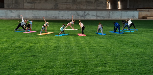 A group of people is practicing yoga on colorful mats spread across a lush green lawn under a concrete structure. The participants are performing a stretching pose and are dressed in casual athletic wear.