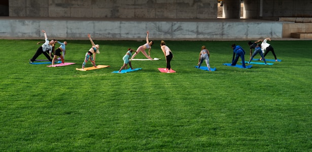 A group of people is practicing yoga on colorful mats spread across a lush green lawn under a concrete structure. The participants are performing a stretching pose and are dressed in casual athletic wear.