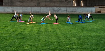 A group of people is practicing yoga on colorful mats spread across a lush green lawn under a concrete structure. The participants are performing a stretching pose and are dressed in casual athletic wear.