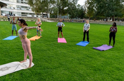 A group of six people are practicing yoga on colorful mats arranged in a line on a lush green lawn. A woman in pink yoga attire is leading the session, standing with arms extended. The participants, displaying various poses, are positioned at a distance from a modern building with large windows. Trees border the area, creating a serene outdoor environment.