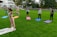 A group of six people are practicing yoga on colorful mats arranged in a line on a lush green lawn. A woman in pink yoga attire is leading the session, standing with arms extended. The participants, displaying various poses, are positioned at a distance from a modern building with large windows. Trees border the area, creating a serene outdoor environment.