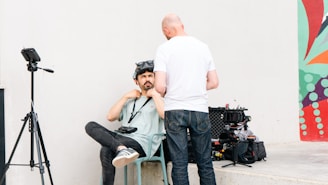Two men are engaged in a conversation near a wall, with one seated and wearing virtual reality goggles, while the other stands facing away. Nearby equipment includes a camera on a tripod and an open case with various video gear. The setting has a modern mural on the wall with abstract patterns.