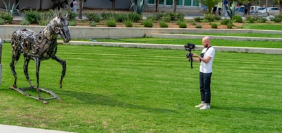 A man wearing a white t-shirt and jeans is operating a camera on a stabilizer while standing on a green lawn. He appears to be filming a sculpture of a horse made from metallic parts, positioned in front of him. The background includes a landscaped area with plants and a building.
