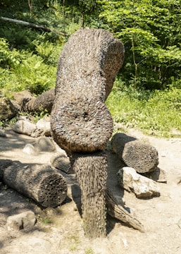 A large sculpture made from numerous coins embedded into logs, forming a unique structure situated outdoors in a wooded area. The sculpture appears to be an artistic installation among a mix of natural materials and lush greenery.