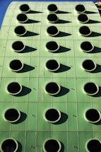 A green building facade with a grid of round, black circular openings that are evenly spaced. The structure's surface appears smooth and metallic, reflecting sunlight and casting shadows from each circular opening.