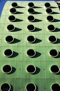 A green building facade with a grid of round, black circular openings that are evenly spaced. The structure's surface appears smooth and metallic, reflecting sunlight and casting shadows from each circular opening.