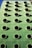 A green building facade with a grid of round, black circular openings that are evenly spaced. The structure's surface appears smooth and metallic, reflecting sunlight and casting shadows from each circular opening.