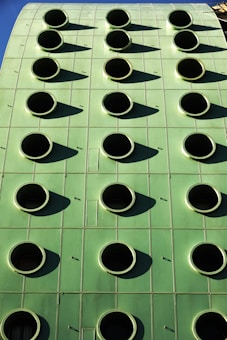 A green building facade with a grid of round, black circular openings that are evenly spaced. The structure's surface appears smooth and metallic, reflecting sunlight and casting shadows from each circular opening.