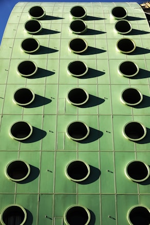 A green building facade with a grid of round, black circular openings that are evenly spaced. The structure's surface appears smooth and metallic, reflecting sunlight and casting shadows from each circular opening.