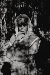 A person with dreadlocks stands in a thoughtful pose, wearing sunglasses and a patterned shirt, amidst dappled sunlight filtering through surrounding foliage.