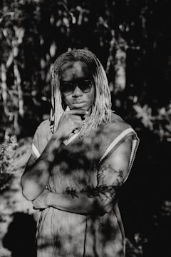 A person with dreadlocks stands in a thoughtful pose, wearing sunglasses and a patterned shirt, amidst dappled sunlight filtering through surrounding foliage.