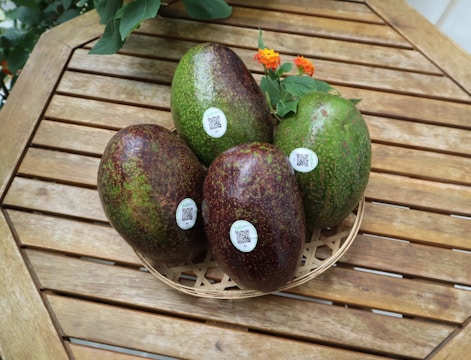 Several avocados with stickers are placed in a woven basket on a wooden table. The table is outdoors and surrounded by a few green leaves with small orange flowers.