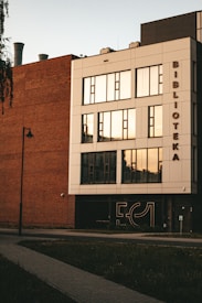 A modern building with a combination of red brick and white facade, featuring large, reflective windows. The word 'BIBLIOTEKA' is vertically displayed on the side. There is a sidewalk and a street lamp in front, with grass areas and a clean sky providing a serene backdrop.