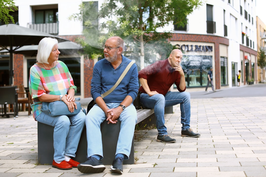 three people sitting on a bench talking to each other, A man smokes a cigarette on a bench and disturbs the elderly coup