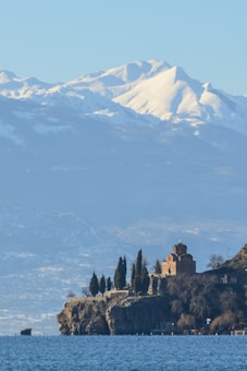 A serene landscape featuring a church located on a cliff overlooking a body of water. The background includes snow-capped mountains under a clear blue sky. Evergreen trees surround the church, adding to the scenic beauty.