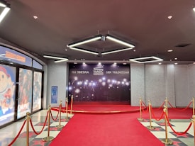 A red carpet leads to a backdrop with bright lights and the words in multiple languages, suggesting a cinematic or award-themed environment. Velvet ropes with gold stanchions line the carpet, and the adjacent wall features movie posters.