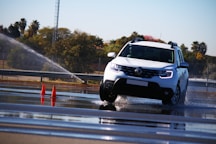 A white car is maneuvering on a wet road surface with water spraying around it. Orange traffic cones are positioned nearby, indicating a possible driving course or test track. Trees and a fence line can be seen in the background under a clear sky.