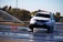 A white car is maneuvering on a wet road surface with water spraying around it. Orange traffic cones are positioned nearby, indicating a possible driving course or test track. Trees and a fence line can be seen in the background under a clear sky.