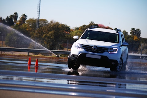 A white car is maneuvering on a wet road surface with water spraying around it. Orange traffic cones are positioned nearby, indicating a possible driving course or test track. Trees and a fence line can be seen in the background under a clear sky.
