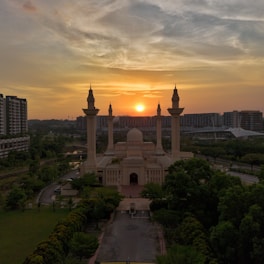 A mosque stands prominently in the foreground with its tall minarets reaching towards a sunset. The sky is filled with a mix of orange and grey hues. Surrounding the mosque are lush green trees and urban buildings in the background.