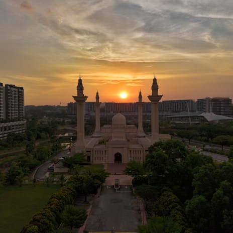 A mosque stands prominently in the foreground with its tall minarets reaching towards a sunset. The sky is filled with a mix of orange and grey hues. Surrounding the mosque are lush green trees and urban buildings in the background.