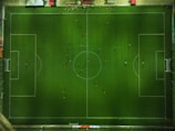Aerial view of a football field with players scattered across the pitch. The field is perfectly marked with white lines, and players are in various positions, casting long shadows. Along the top, there is a line of spectators or team benches on the sidelines.
