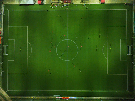 Aerial view of a football field with players scattered across the pitch. The field is perfectly marked with white lines, and players are in various positions, casting long shadows. Along the top, there is a line of spectators or team benches on the sidelines.