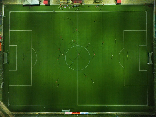 Aerial view of a football field with players scattered across the pitch. The field is perfectly marked with white lines, and players are in various positions, casting long shadows. Along the top, there is a line of spectators or team benches on the sidelines.