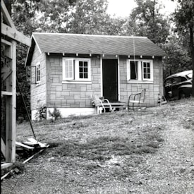A small shed or cabin with a gabled roof and a facade resembling masonry siding. It has a front door flanked by two windows and two metal chairs are placed on its small porch. A vintage car is partially visible to the right, near the structure. Surrounding the building are trees and a grassy yard with a gentle slope.