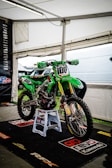 A green motocross bike is displayed on a stand inside a white tent. The bike has various sponsorship logos on its body, including Motul, Pro Circuit, and Kawasaki. It is supported by a blue rev stand and sits on a branded black mat.