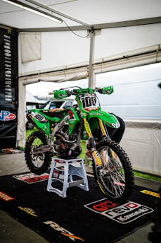 A green motocross bike is displayed on a stand inside a white tent. The bike has various sponsorship logos on its body, including Motul, Pro Circuit, and Kawasaki. It is supported by a blue rev stand and sits on a branded black mat.