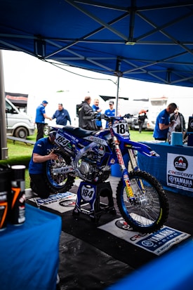 A motocross bike with the number 184 is displayed under a blue tent, surrounded by people wearing blue attire. The bike is mounted on a stand and is being worked on by a technician. Various equipment and banners can be seen around, including those with Yamaha branding.