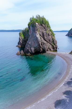 A picturesque coastal scene featuring a tall, rocky island with lush green vegetation on top. The island is surrounded by clear turquoise water that gently laps against a narrow, crescent-shaped sandy beach. The sky is pale blue with a few scattered clouds, and the horizon stretches into the distance.